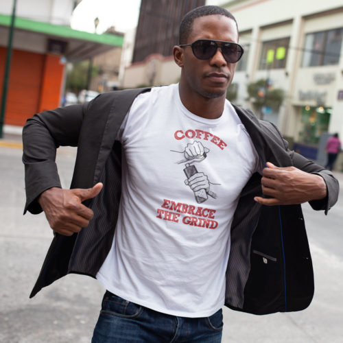 A man in the street putting on a jacket wearing a white t-shirt with a drawing of hands using a handheld coffee grinder with the words COFFEE EMBRACE THE GRIND in red lettering. Designed by Kenny Velez