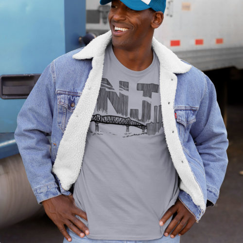 A man wearing a slate gray t-shirt with a line drawing of the Pulaski Skyway and Kearny power plant with the initials NJ. Designed by Kenny Velez.