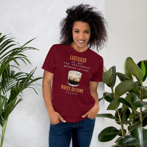 Woman wearing a cranberry red t-shirt with the words Caucasian is not the preferred nomenclature. White Russian please. A play on quotes and themes from the movie The Big Lebowski. Designed by Kenny Velez