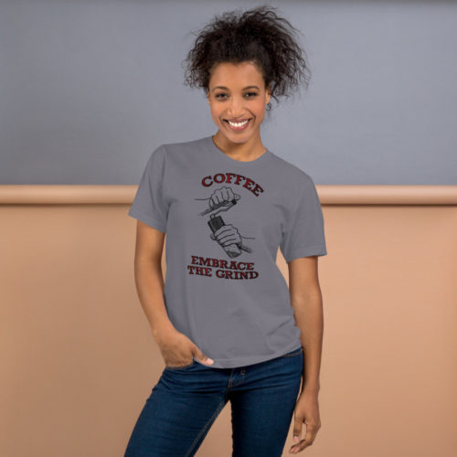 A woman wearing a slate gray t-shirt with a drawing of hands using a handheld coffee grinder with the words COFFEE EMBRACE THE GRIND in red lettering. Designed by Kenny Velez