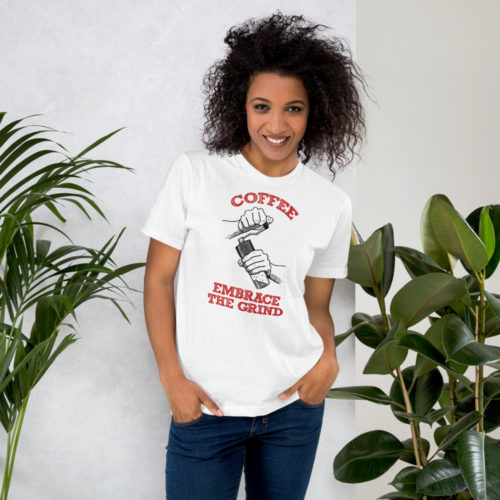 A woman wearing a white t-shirt with a drawing of hands using a handheld coffee grinder with the words COFFEE EMBRACE THE GRIND in red lettering. Designed by Kenny Velez