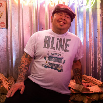 A guy wearing a white t-shirt with a drawing of a Bergenline jitney dollar bus from Union City, NJ with word BLine on top. Designed by Kenny Velez