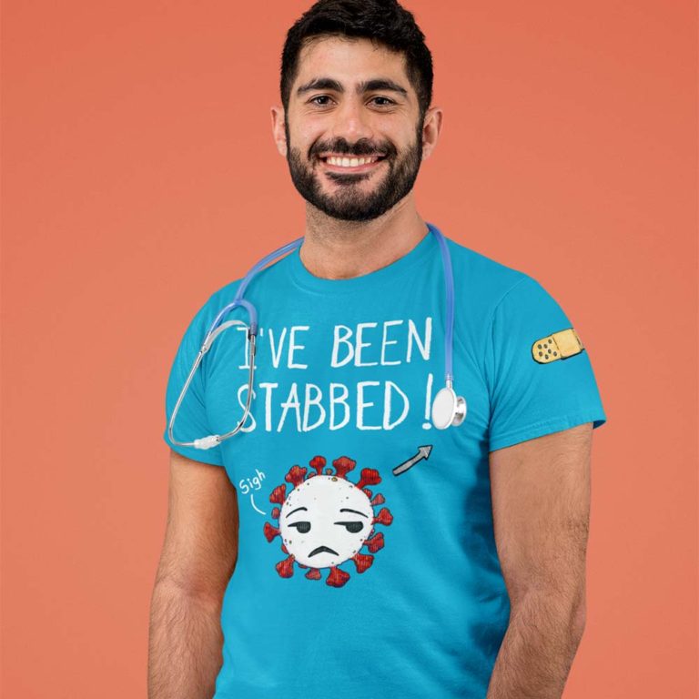 A male healthcare worker wearing a teal blue t-shirt with a drawing of a sighing Covid-19 Coronavirus and the words IVE BEEN STABBED! An arrow next to the drawing points to left sleeve shoulder which has a bandaid. Designed by Kenny Velez.