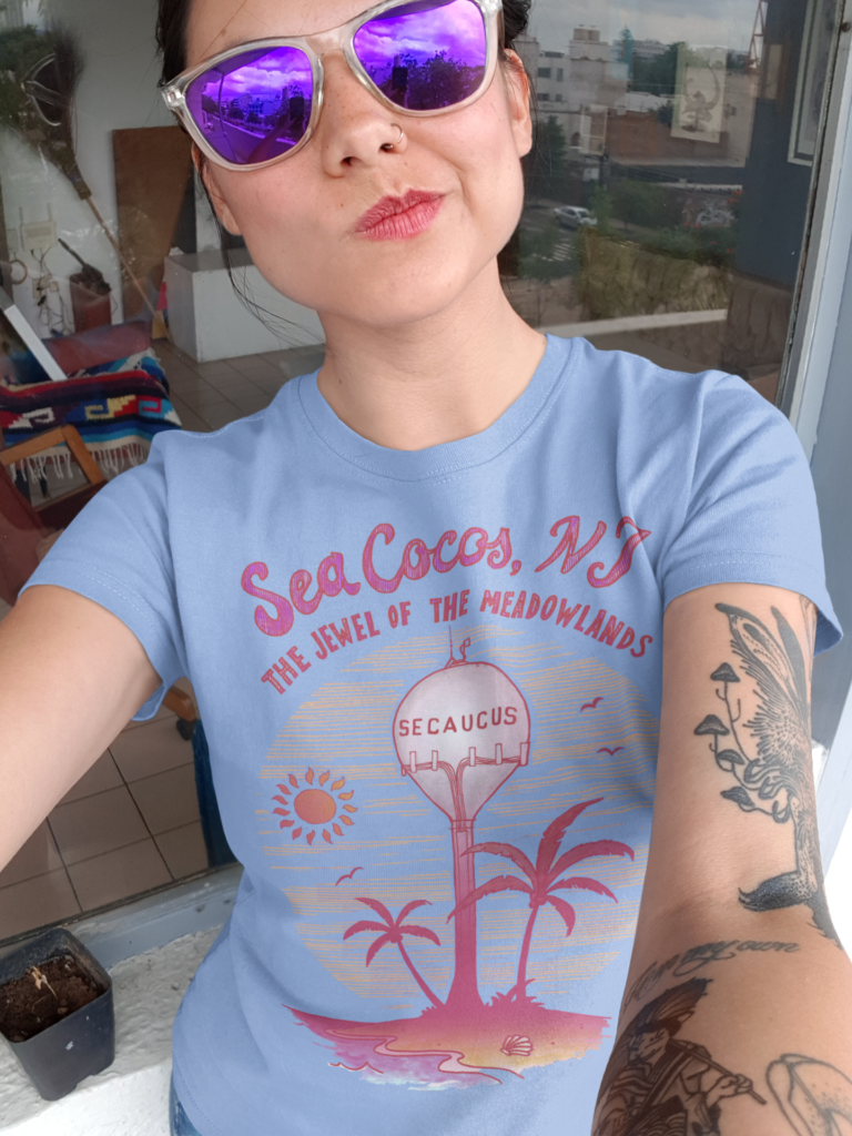 A girl wearing a white t-shirt with a drawing of the water tower from Secaucus, NJ on a beach, with the words Sea Cocos, The Jewel of the Meadowlands. Designed by Kenny Velez.