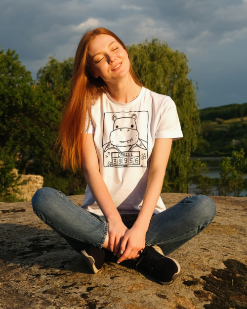 A girl sitting on the ground wearing a t-shirt with a drawing of a mugshot featuring a cartoon hippo in a Hawaiian shirt in the vein of the iconic mugshot of Pablo Escobar. Designed by Kenny Velez.