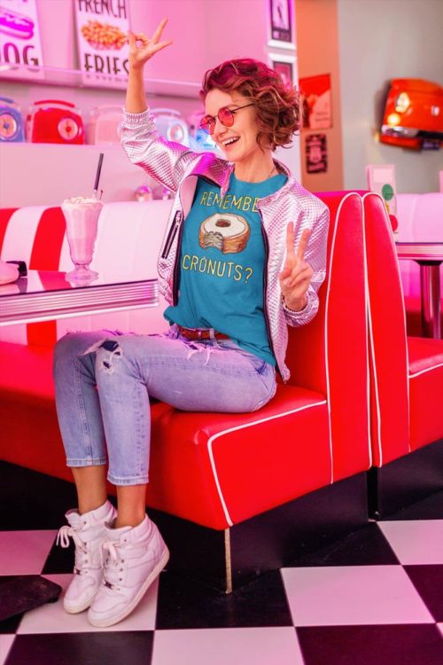 A woman sitting in a classic diner wearing a teal blue t-shirt with a drawing of a bitten cronut with the words REMEMBER CRONUTS? in yellow lettering. Designed by Kenny Velez.