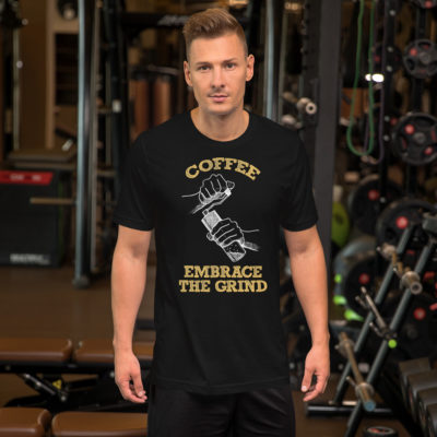 a guy wearing A black t-shirt with a drawing of hands using a handheld coffee grinder with the words COFFEE EMBRACE THE GRIND in yellow lettering. Designed by Kenny Velez