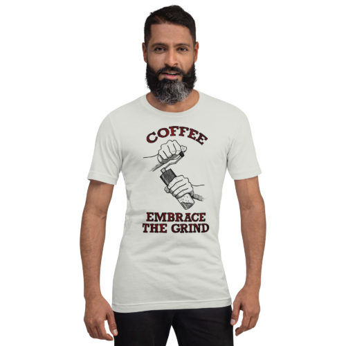 A guy wearing a silver t-shirt with a drawing of hands using a handheld coffee grinder with the words COFFEE EMBRACE THE GRIND in red and black lettering. Designed by Kenny Velez