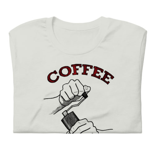 A silver t-shirt with a drawing of hands using a handheld coffee grinder with the words COFFEE EMBRACE THE GRIND in red and black lettering. Designed by Kenny Velez