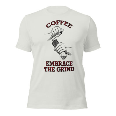 A silver t-shirt with a drawing of hands using a handheld coffee grinder with the words COFFEE EMBRACE THE GRIND in red and black lettering. Designed by Kenny Velez