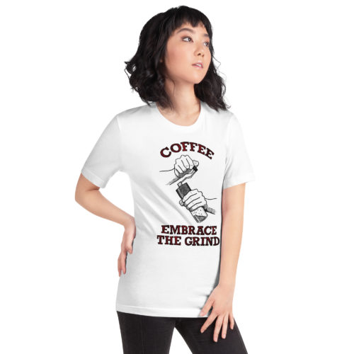 A girl wearing a white t-shirt with a drawing of hands using a handheld coffee grinder with the words COFFEE EMBRACE THE GRIND in red and black lettering. Designed by Kenny Velez